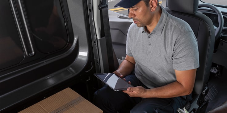 A man in a parked Transit® Cargo Van with driver seat facing cargo area working on a tablet