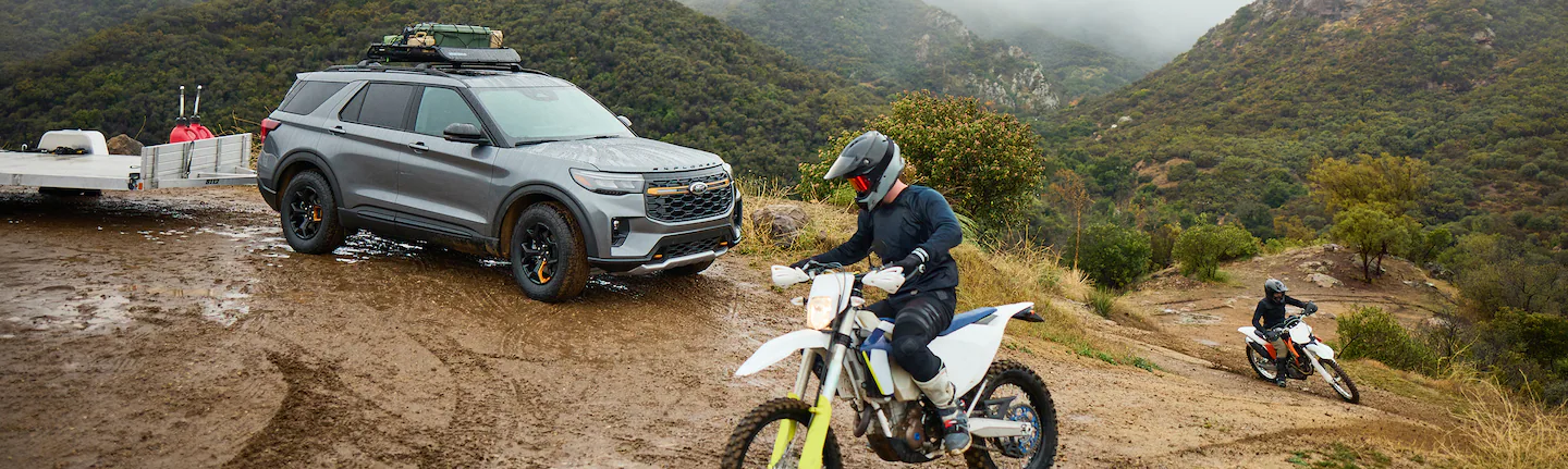 Grey Ford SUV towing a small utility trailer on a muddy trail, with a dirt bike in the foreground and forested hills in the background