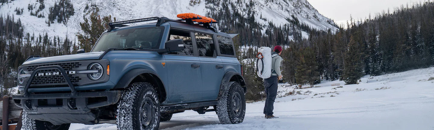 A person with camping gear stands behind a Ford Bronco parked on a snow-covered, tree-lined slope.
