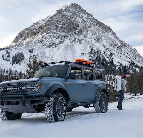 A person with camping gear stands behind a Ford Bronco parked on a snow-covered, tree-lined slope.