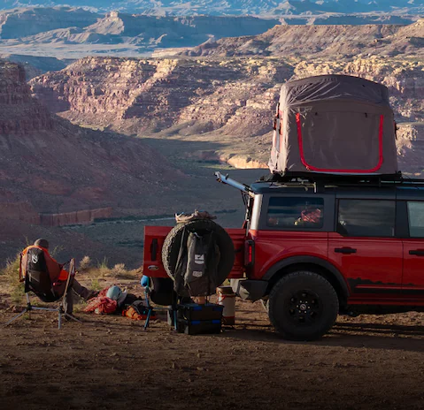 Red Ford Bronco with rooftop tent in a canyon, a person sits in a chair near camping gear.