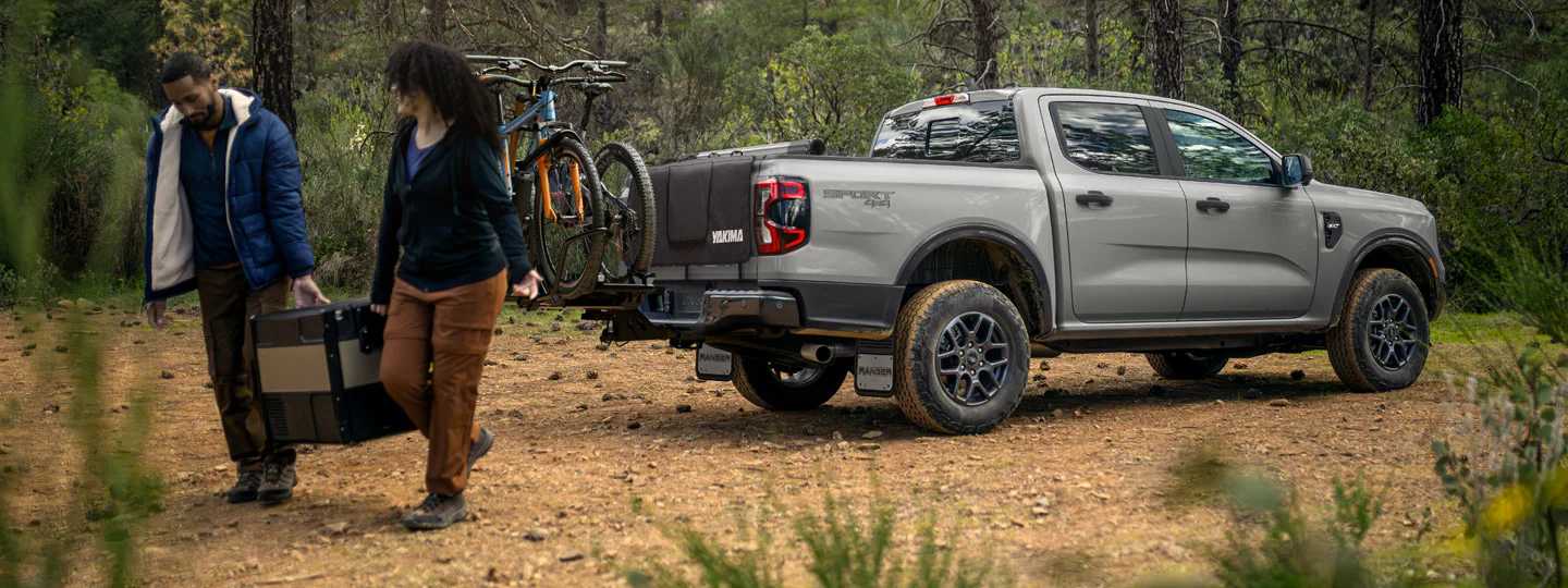 A man and a woman carry camping gear away from their 2026 Ford Ranger® XLT parked on a rocky dirt path surrounded by pine trees