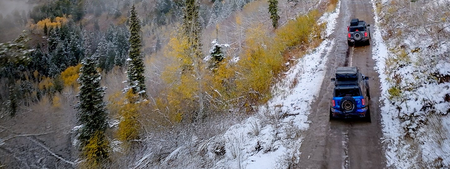 A Ford Bronco SUV, with available roof rack, is parked on a snowy road, with a forest in the background