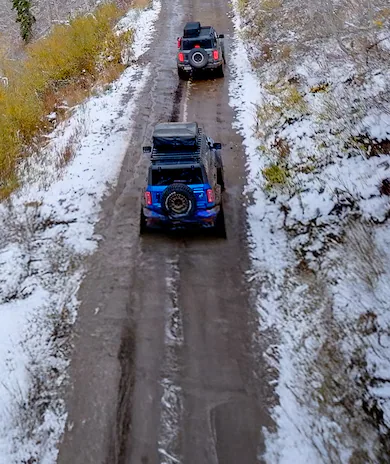 A Ford Bronco SUV, with available roof rack, is parked on a snowy road, with a forest in the background