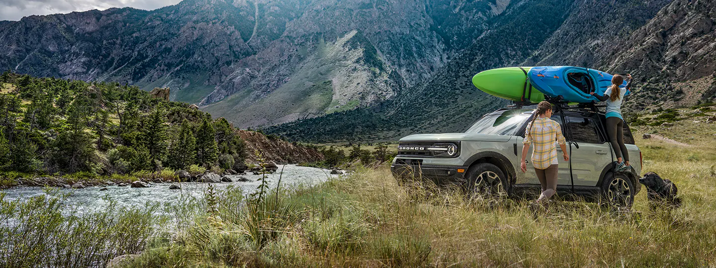 A 2025 Bronco® next to river with people handling kayaks on roof rack