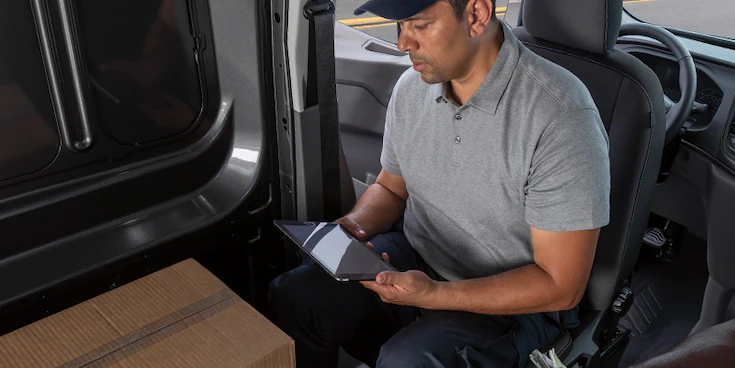 A man in a parked Transit® Cargo Van with driver seat facing cargo area working on a tablet